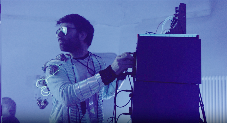 The photo shows an electronic musician in retro-chic outfits playing an analog sampler