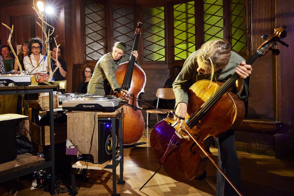 we see a contra bassist and a cellist performing while pulling an elastng band that is attached to the turntable and moves the platter