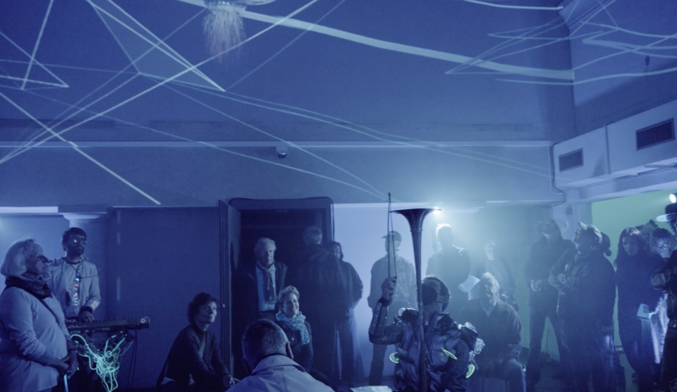 Two time travelers in the time machine, a space with much video, fog machine, they wear funny costumes and are playing self made instruments or synthesizers